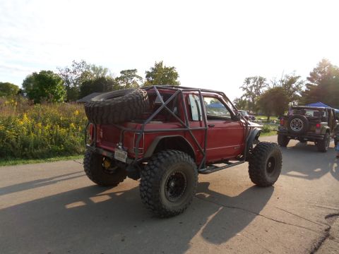1989 Ford Bronco - A heavily modified red off-road vehicle with an extensive external roll cage and a spare tire mounted on the rear is parked on a paved surf…
