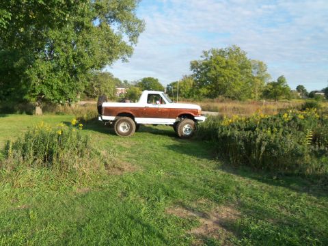 1989 Ford Bronco - This lifted Ford Bronco is parked on a grassy area with trees and vegetation in the background under a partly cloudy sky.