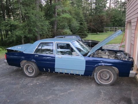 1991 Ford LTD Crown Victoria - A two-tone blue 1991 Ford Crown Victoria LTD is parked outside a garage with its hood open, revealing the engine compartment.