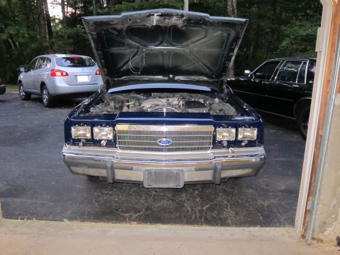 1991 Ford LTD Crown Victoria - The front of a blue Ford Crown Victoria with its hood open, revealing the engine bay. A silver SUV and a black sedan are visible in the bac…