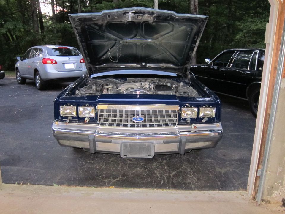 Blue Ford Crown Victoria with hood open, showing engine. Another car is parked behind it
