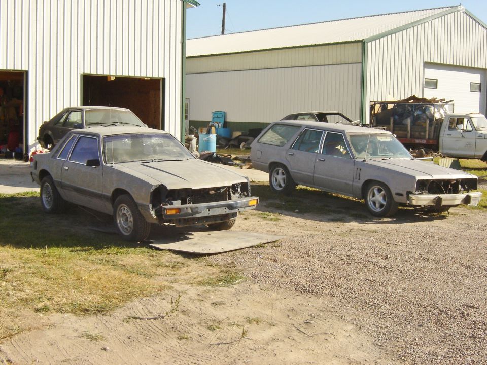 Two dusty cars, a sedan and a station wagon, parked outside a garage. The sedan is in the foreground, missing its grille and headlights. The station wagon is to the right, also appearing weathered