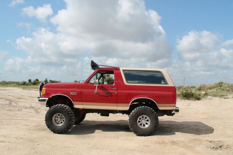 1990 Ford Bronco - This lifted Ford Bronco, likely from the late 1980s, sits on a sandy terrain under a cloudy sky.
