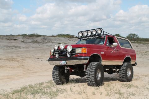1990 Ford Bronco - A modified 1990 Ford Bronco sits on a sandy terrain under a cloudy sky, equipped with numerous auxiliary lights.