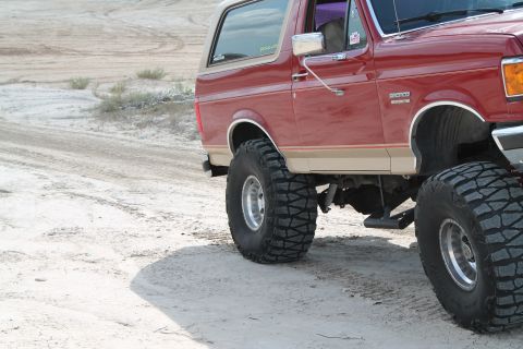 1990 Ford Bronco - A lifted red 1987 Ford Bronco with tan trim drives on a sandy, off-road terrain.