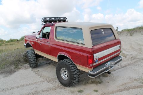 1990 Ford Bronco - This lifted 1990 Ford Bronco features a tan removable top and a roof-mounted light bar, seen off-road on a sandy dune.