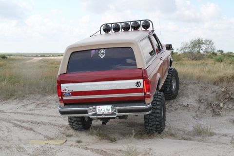 1990 Ford Bronco - Rear view of a lifted 1990 Ford Bronco with large tires, driving on a dirt trail.
