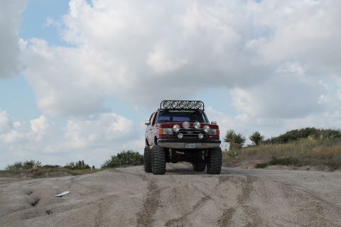 1990 Ford Bronco - A modified red Ford Bronco navigates a sandy terrain under a cloudy sky.
