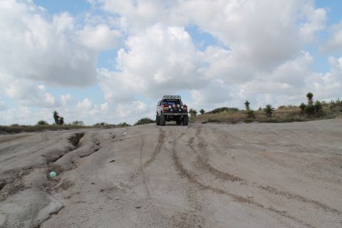 1990 Ford Bronco - A red 1990 Ford Bronco with off-road modifications navigates a sandy incline under a cloudy sky.