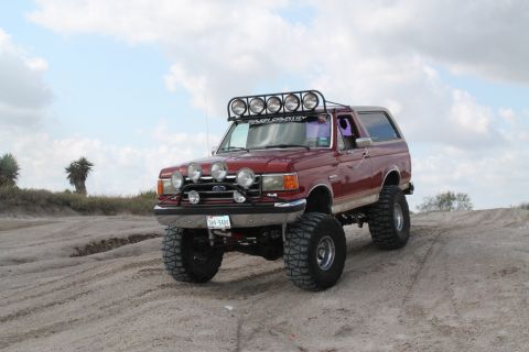 1990 Ford Bronco - A modified Ford Bronco drives on a sandy terrain under a cloudy sky.