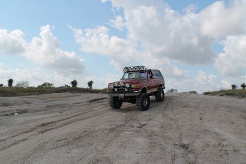 1990 Ford Bronco - 1990 Ford Bronco with off-road modifications navigating a sandy terrain under a cloudy sky.
