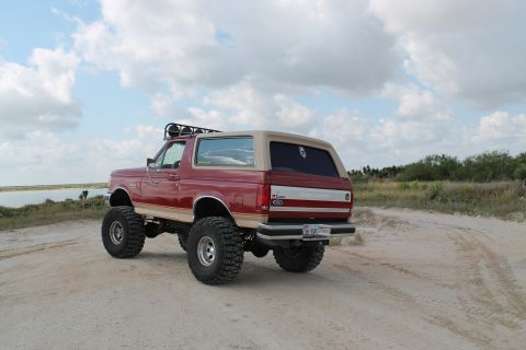 1990 Ford Bronco - This lifted 1990 Ford Bronco is shown from the rear on a sandy track with a body of water in the background.