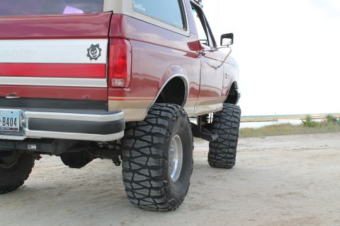 1990 Ford Bronco - Rear view of a lifted 1990 Ford Bronco with oversized tires, parked on a sandy surface near water.