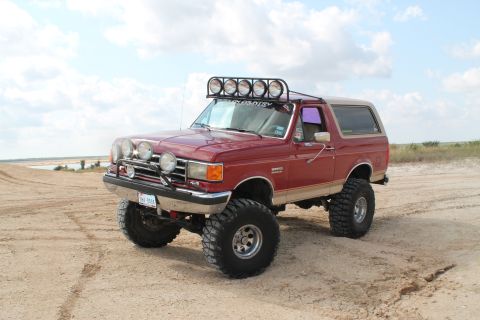 1990 Ford Bronco - Modified 1990 Ford Bronco with extra lights and large tires parked on a sandy area.