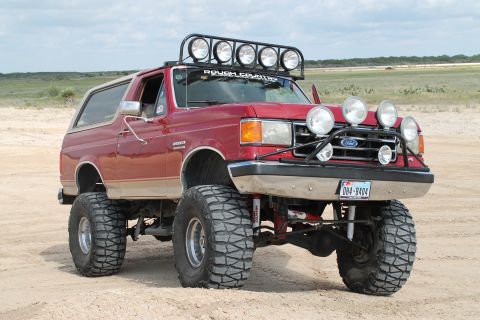 1990 Ford Bronco - This modified Ford Bronco, likely from the late 1980s, sits on a sandy terrain under a cloudy sky.