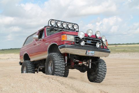 1990 Ford Bronco - Modified 1990 Ford Bronco with off-road lights and tires sits on sandy terrain.