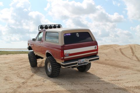 1990 Ford Bronco - This modified Ford Bronco is seen from the rear on a sandy terrain under a cloudy sky.