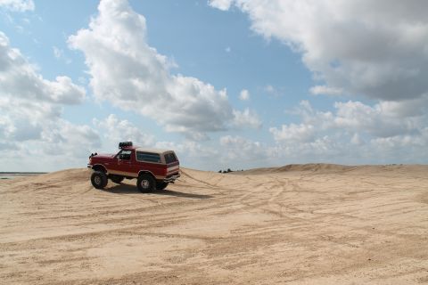 1990 Ford Bronco - A red 1990 Ford Bronco navigates sandy terrain under a partly cloudy sky.