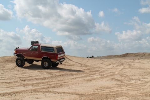 1990 Ford Bronco - Red 1990 Ford Bronco with off-road modifications in a sandy landscape under a cloudy sky.