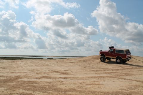 1990 Ford Bronco - A red Ford Bronco with a camper shell navigates sandy terrain under a partly cloudy sky.