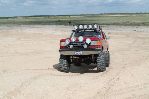 1990 Ford Bronco - Modified 1990 Ford Bronco with extensive lighting and large tires sits on a sandy terrain.