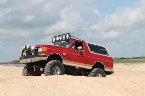 1990 Ford Bronco - A modified 1990 Ford Bronco with off-road lights is partially submerged in sand under a cloudy sky.