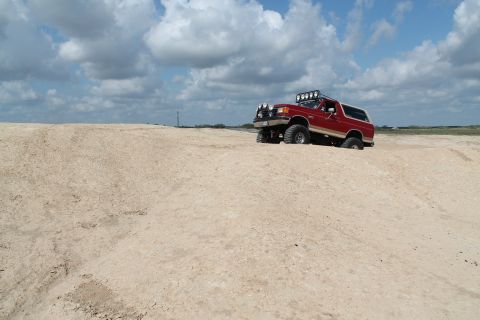 1990 Ford Bronco - Red 1990 Ford Bronco navigating a sandy terrain under a cloudy sky.