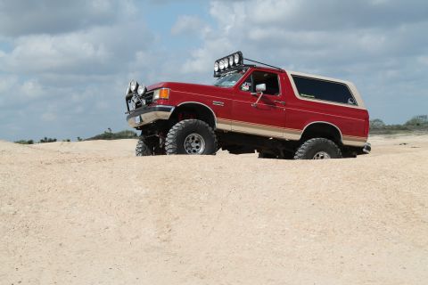 1990 Ford Bronco - A lifted red Ford Bronco navigates sandy terrain under a cloudy sky.