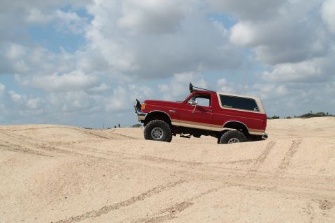 1990 Ford Bronco - A red 1990 Ford Bronco navigates sandy terrain under a cloudy sky.