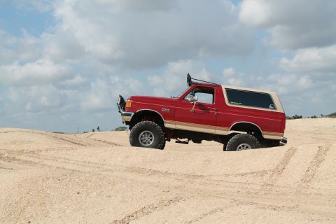 1990 Ford Bronco - Red 1990 Ford Bronco navigating sandy terrain with a lifted suspension and large off-road tires.