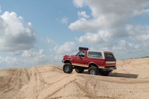 1990 Ford Bronco - This lifted Ford Bronco navigates sandy terrain under a bright, cloudy sky.