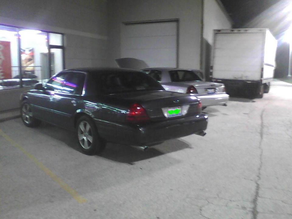 Dark sedan parked in a lot at night, with a silver sedan and a box truck behind it