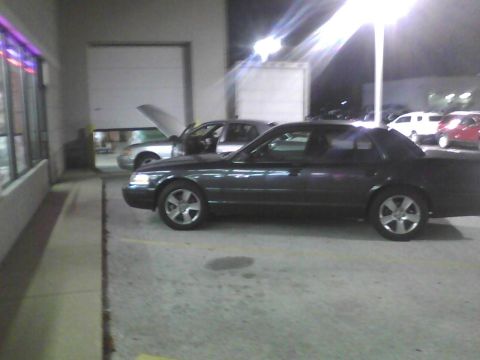 2003 Ford Crown Victoria - A dark 2003 Ford Crown Victoria is parked next to another car with its hood up.