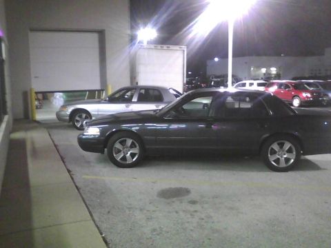 2003 Ford Crown Victoria - A dark 2003 Ford Crown Victoria is parked beside a silver sedan in a lot at night.