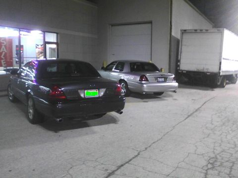 2003 Ford Crown Victoria - Two Ford Crown Victorias are parked next to each other outside a commercial building with a box truck nearby.