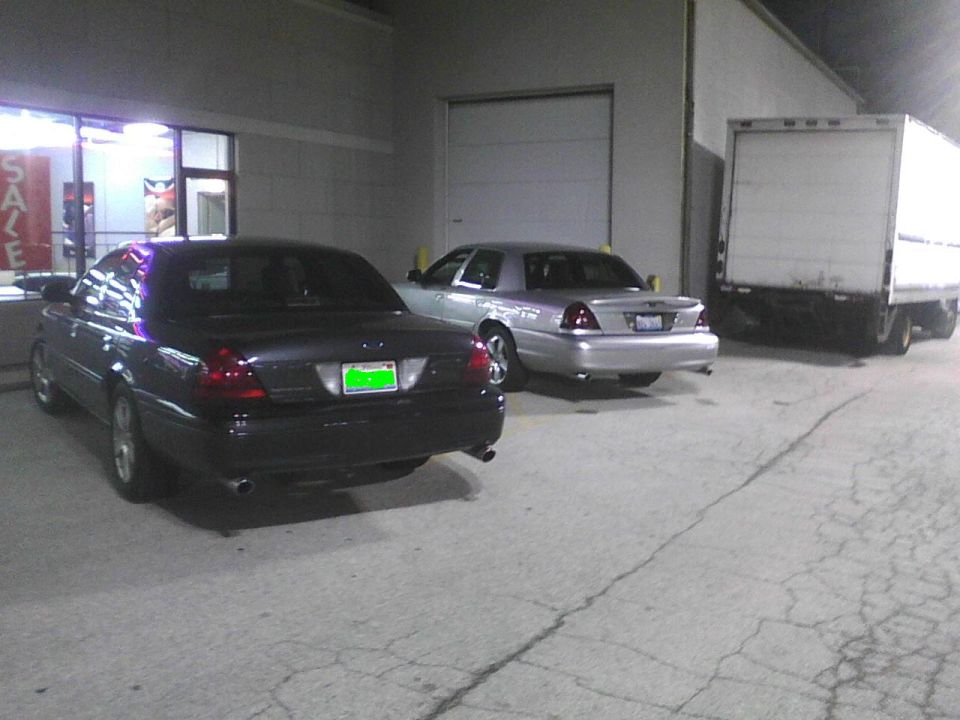 Two Ford Crown Victorias parked outside a building, one dark and one silver, with a box truck to the right