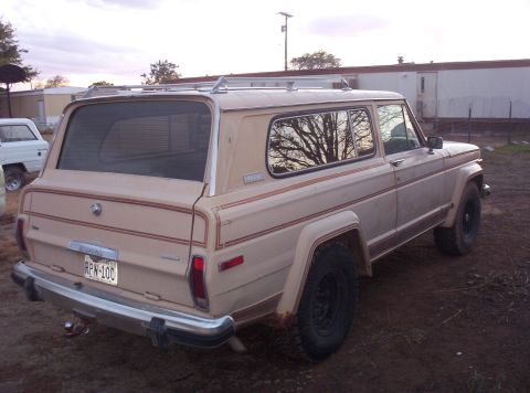 1988 Ford Bronco - A tan Jeep Wagoneer is parked on dirt ground with a white vehicle in the background.