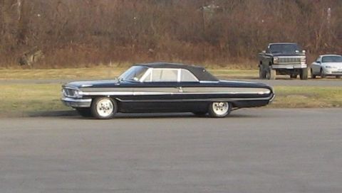1964 Ford Galaxie - A black convertible with chrome trim is parked on the side of the road, with other vehicles in the distance.