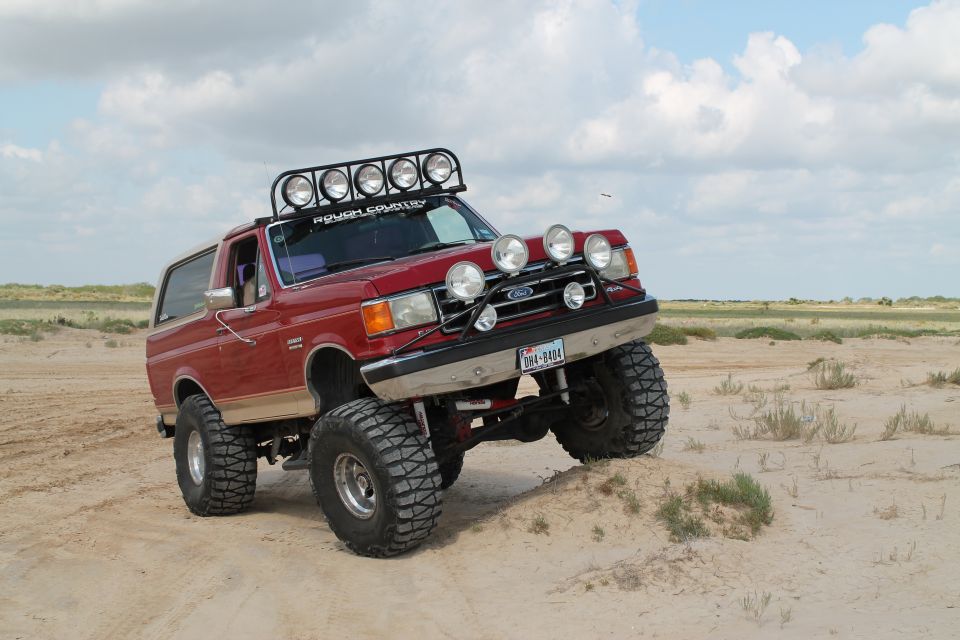 Red 1990 Ford Bronco with off-road lights and roof rack on sandy terrain