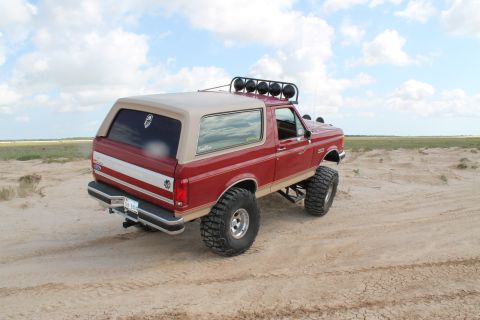 1990 Ford Bronco - A lifted 1990 Ford Bronco with a tan removable top drives on a sandy landscape under a cloudy sky.