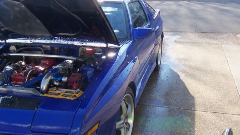 1989 Mitsubishi Starion - The engine bay of a blue sports car is visible with the hood up, showing various engine parts.