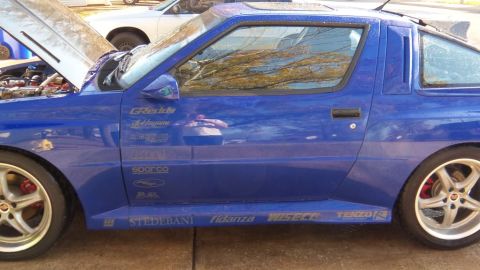 1989 Mitsubishi Starion - Side view of a blue 1989 Mitsubishi Starion, showing racing decals and an open hood.