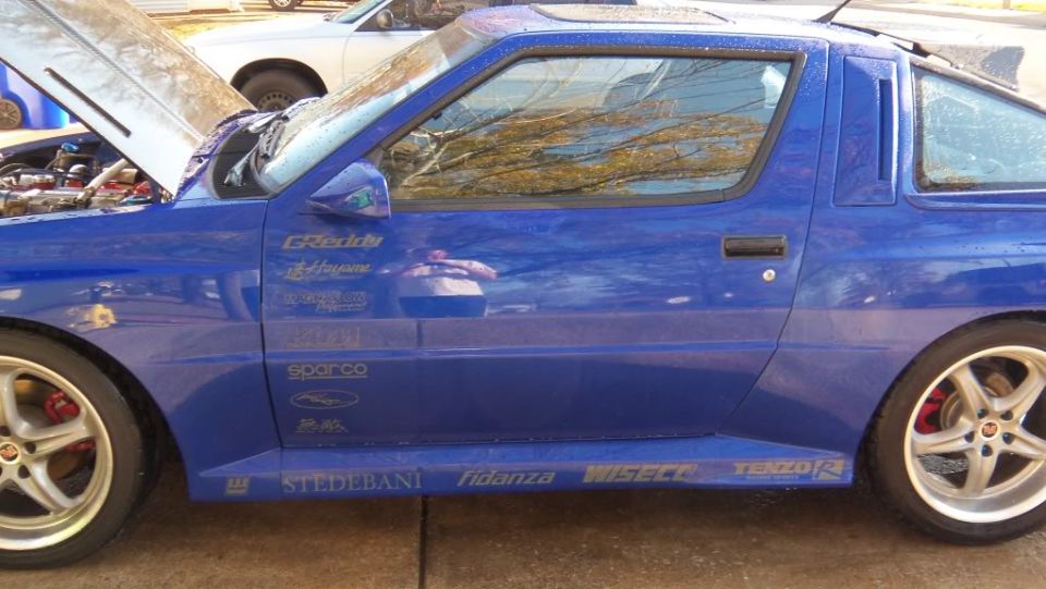 Blue 1989 Mitsubishi Starion with racing decals on the side, hood open, another car in background