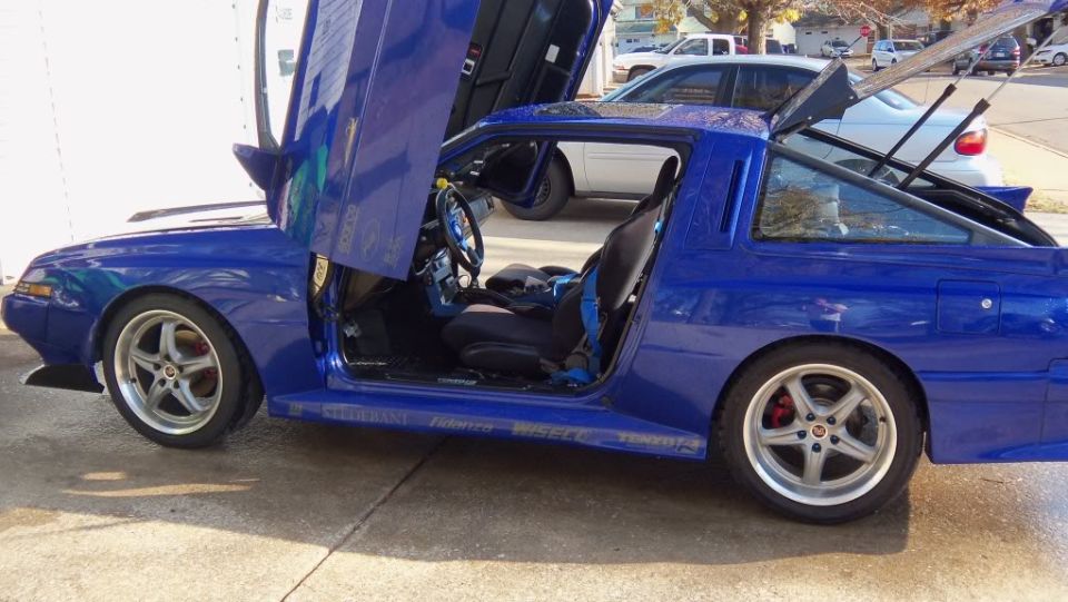 1989 Mitsubishi Starion with custom blue paint and lambo doors open, showing racing seats and roll cage. Other cars visible in background