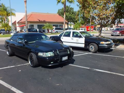 2003 Ford Crown Victoria - Two Panther-platform sedans angled in a parking lot, one civilian black, one LAPD livery.