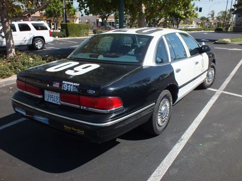 2003 Ford Crown Victoria - Rear quarter of a black-and-white police-themed Ford sedan marked 09 with a 911 plate frame.