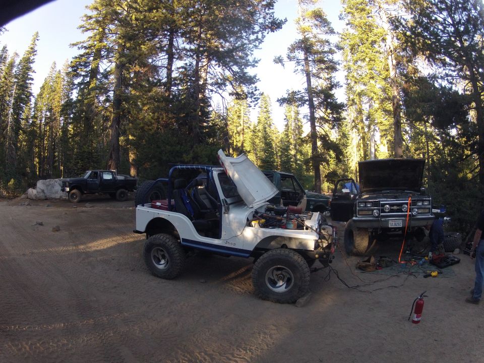 Several off-road vehicles, including a white Jeep and a black pickup truck, are parked in a wooded area with their hoods open, suggesting mechanical work is underway