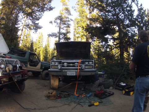 1989 Ford Bronco - A group works on a Chevrolet truck in a forest clearing, with other off-road vehicles nearby.
