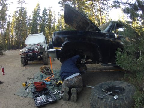 1989 Ford Bronco - A person works on the undercarriage of a black pickup truck with its hood open, while a white Jeep sits nearby in a wooded area.