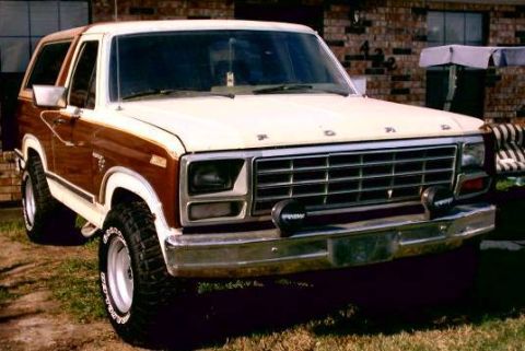 1980 Ford Bronco - Front 3/4 view of a brown and white two-tone full-size Ford Bronco with a white removable hardtop, parked on grass in front of a brick hous…